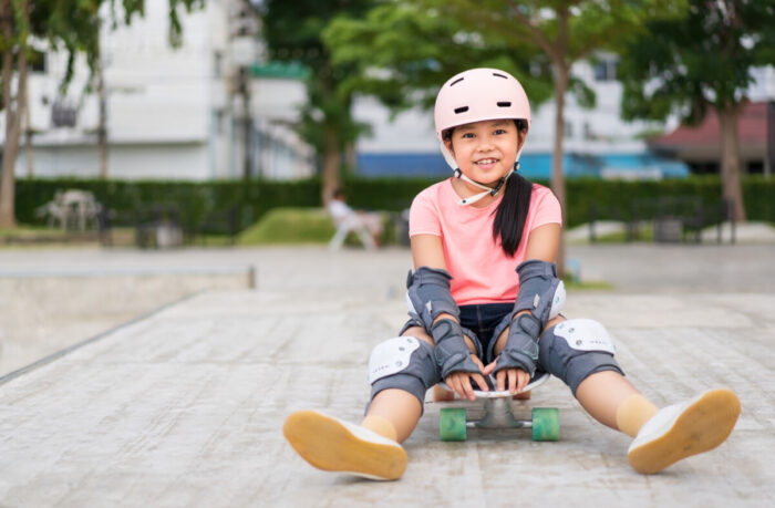 Kleines Mädchen sitzt auf einem Skateboard auf einem asphaltierten Platz, geschützt mit Ellenbogen- und Knieschonern